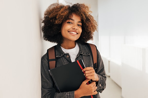 Une jeune femme avec des cheveux bouclés qui portent une veste en jean et tiennent un cahier. Étudiante ou apprenantes, elle semble être souriante et dégage une ambiance positive.